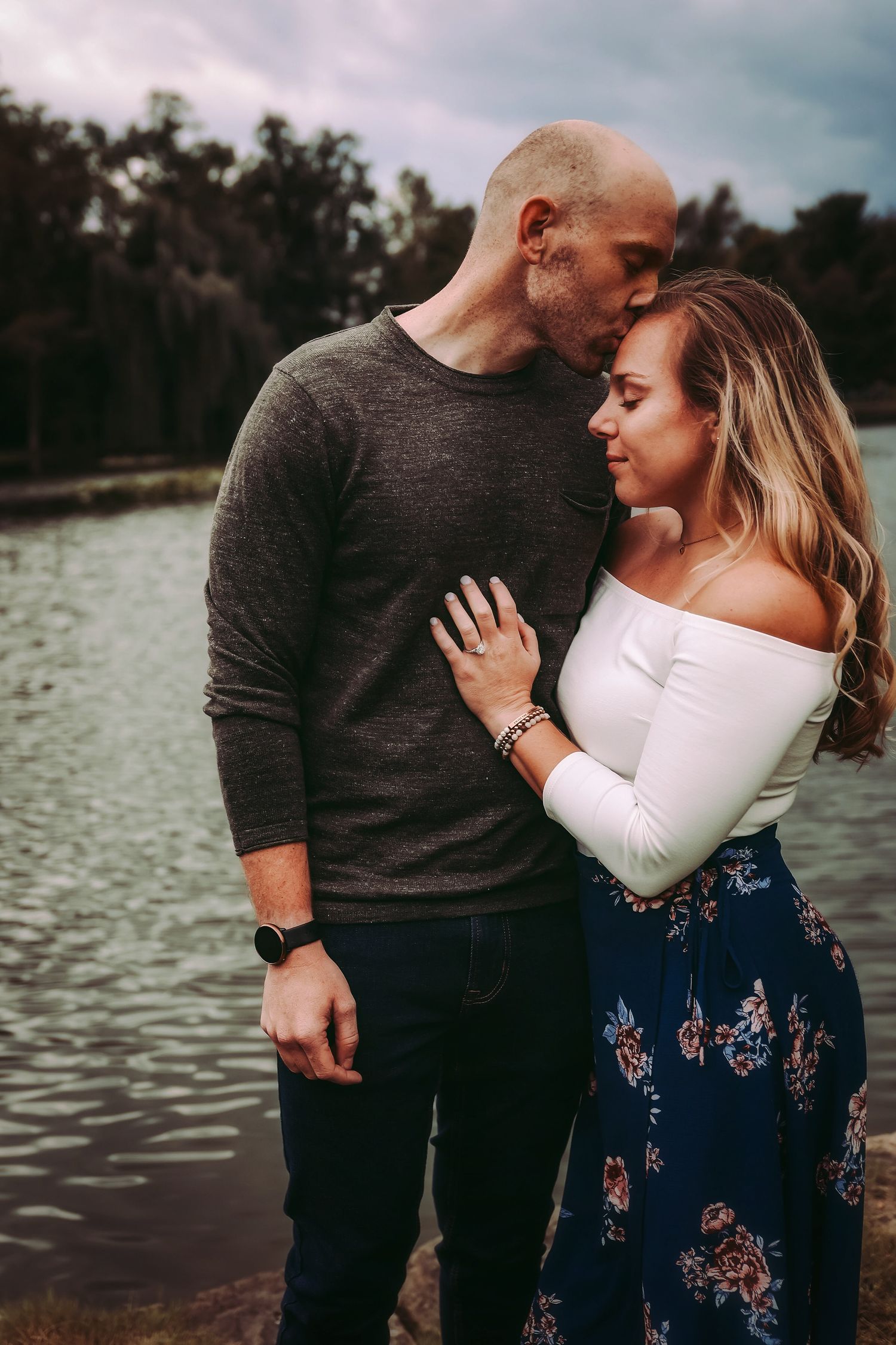 A couple embracing by a lake at sunset with mountains in the background.