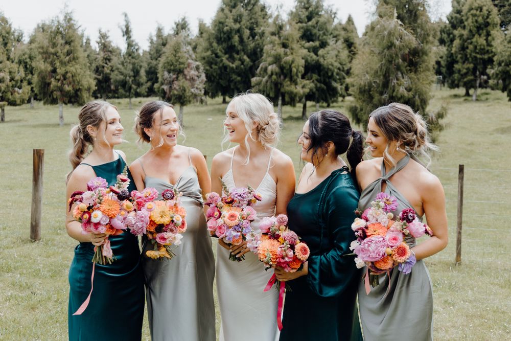 Bridal party in teal and grey dresses hold vibrant coral bouquets while laughing together.