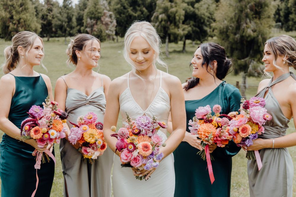 Wedding party members admire their colorful bouquets in natural outdoor setting.