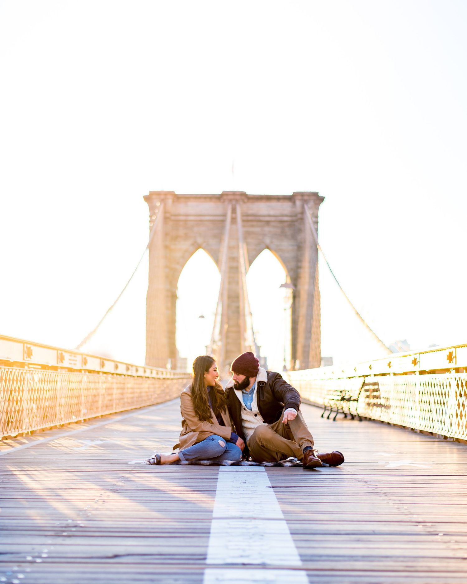 engaged couple sitting in the middle of an empty Brooklyn Bridge at sunrise in NYC while laughing and cuddling