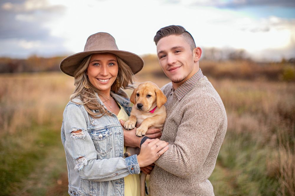 Woman and hat with boyfriend and puppy in a field for a family photo session