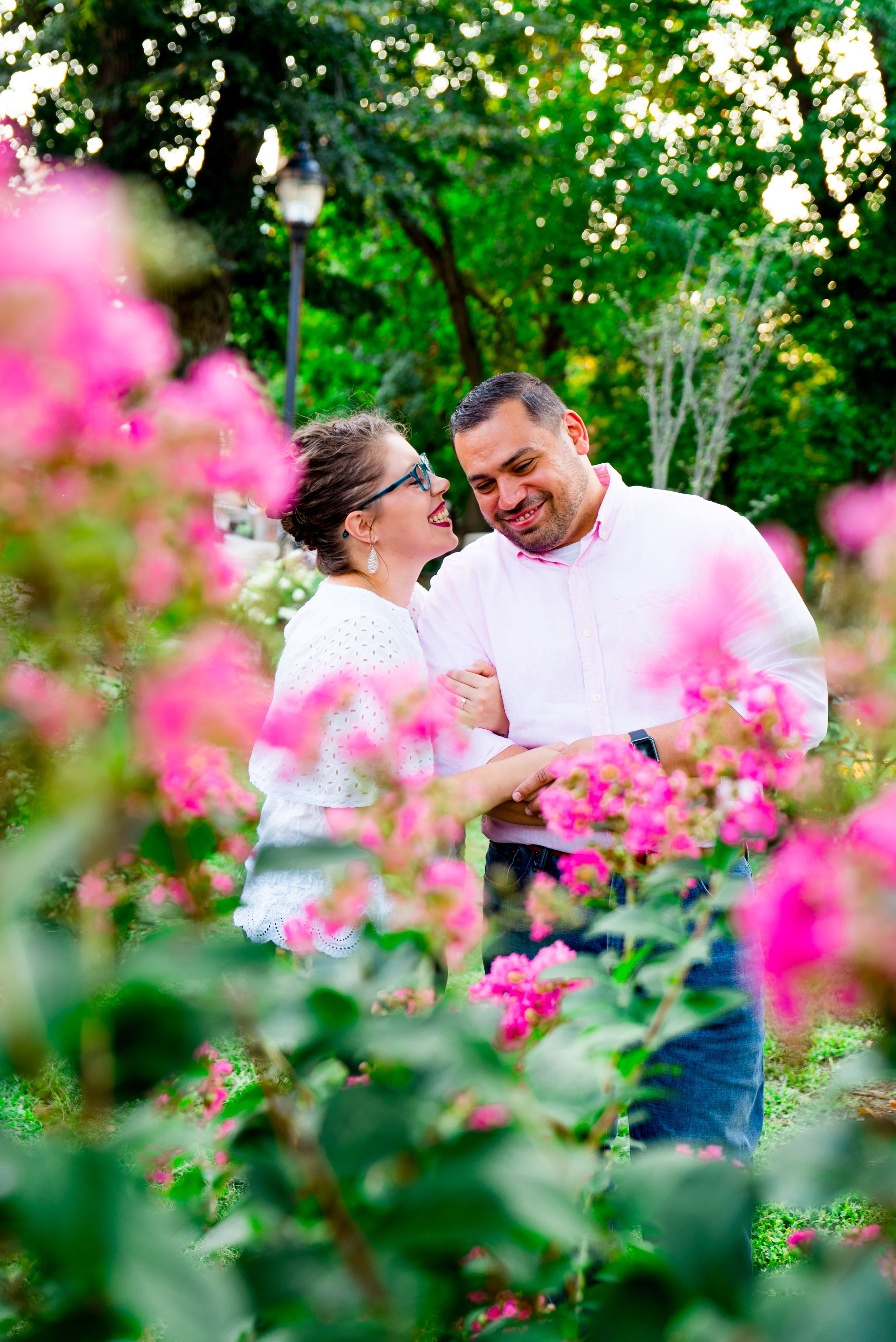 woman in white shirt and red lipstick pulls fiance in close to whisper in his ear and laughs behind a pink flower bush