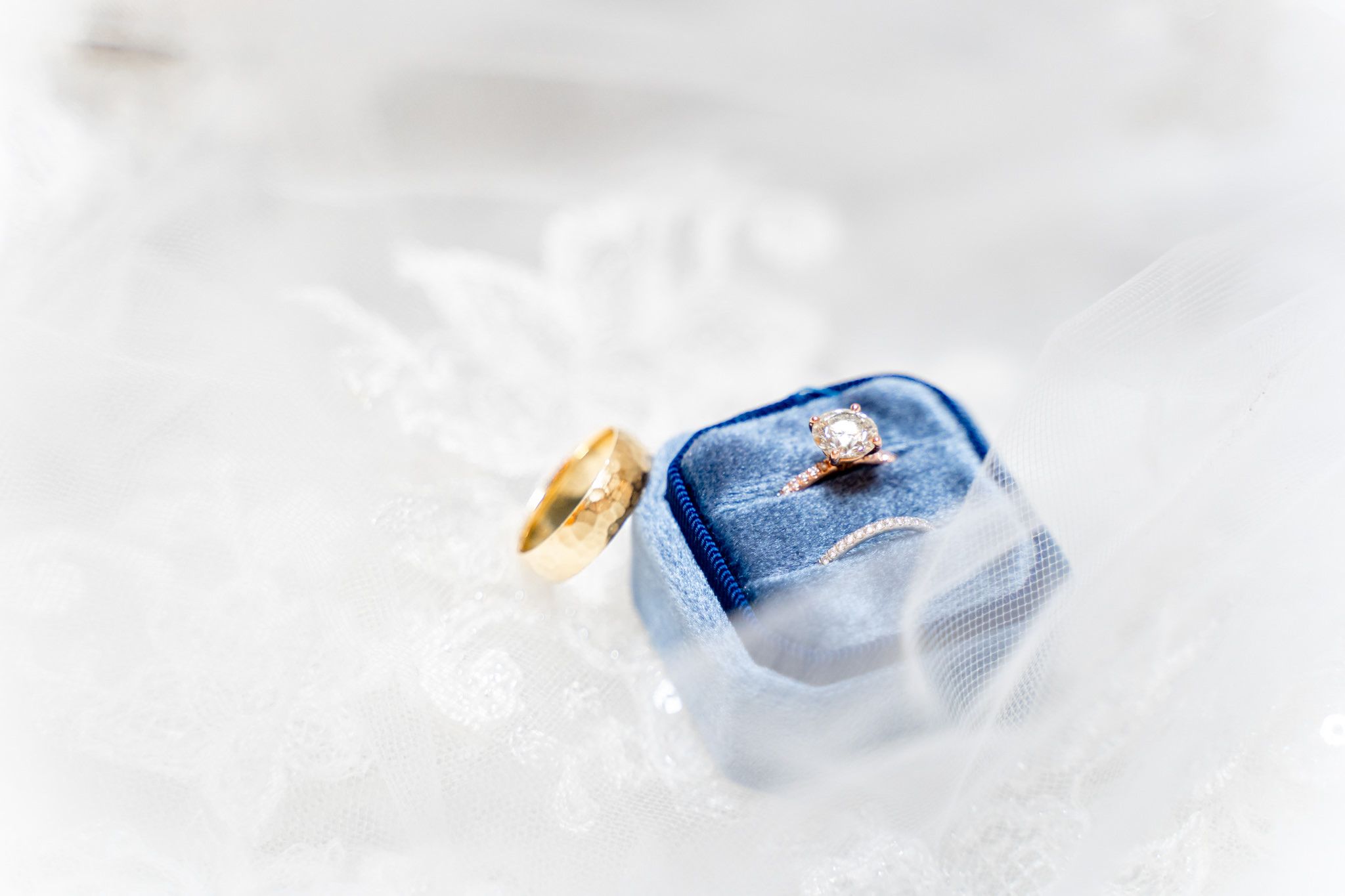 round diamond engagement ring with wedding bands in blue velvet ring box on lace wedding dress