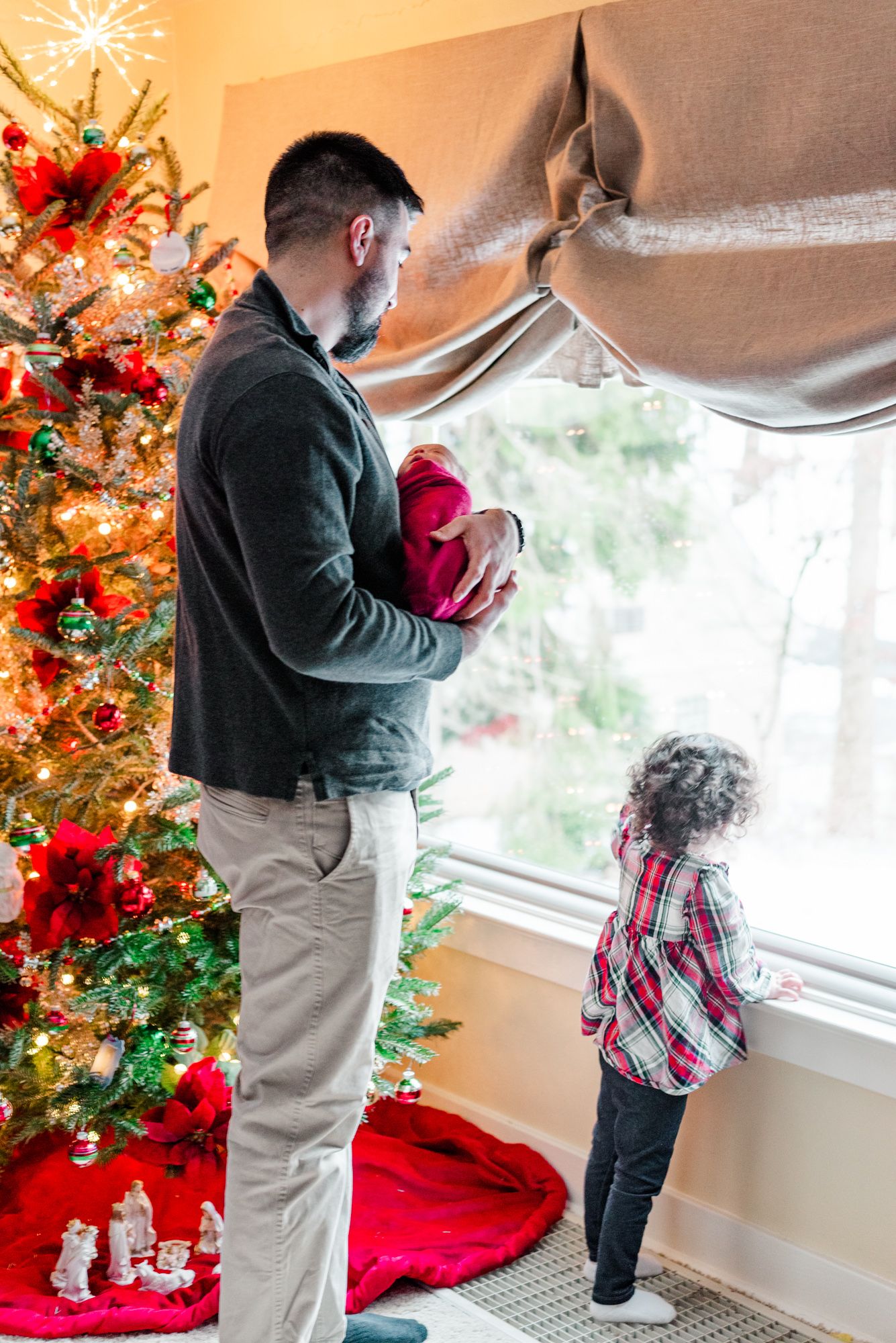 Lifestyle father and big sister holding newborn baby in pink wrap in front of Christmas tree in Pittsburgh, Pennsylvania