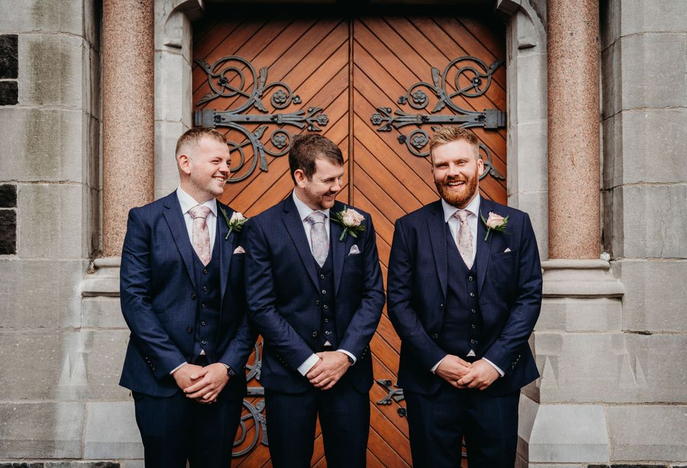 Groom and his two groomsmen at either side of him, outside St Peter's Church Lurgan, in their navy suits and laughing.