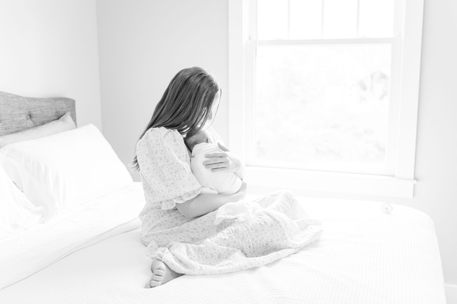 Black and white image of a new mother embracing her newborn sitting on a bed in front of a bright window.