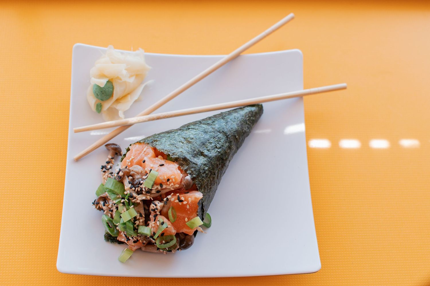 Fresh temaki hand roll with chopsticks served on white square plate against orange background.