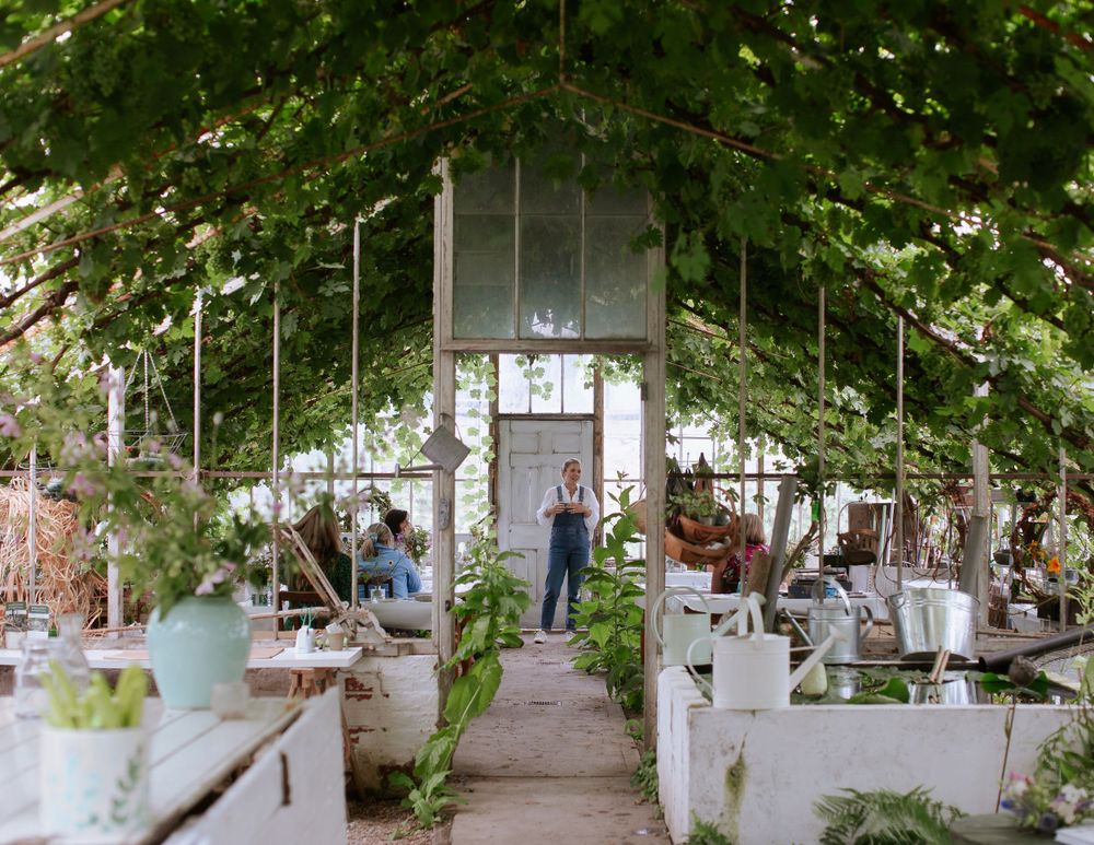 A woman wearing blue dungarees teaches an art class inside a large Victorian greenhouse