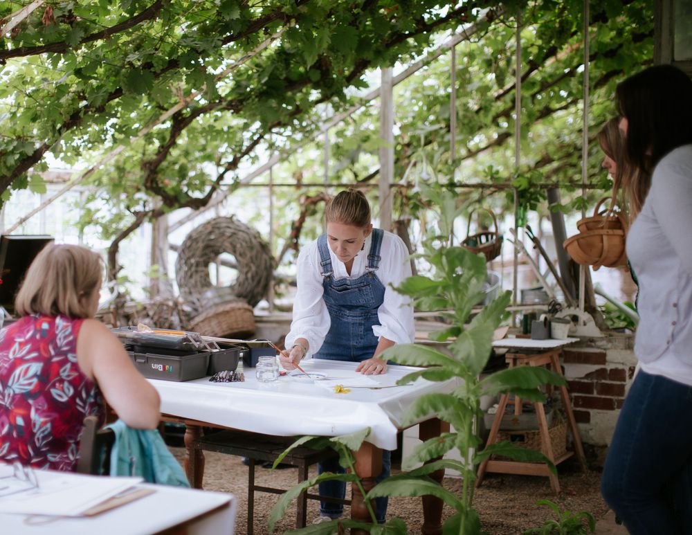 A woman wearing blue dungarees teaches an art class inside a large Victorian greenhouse