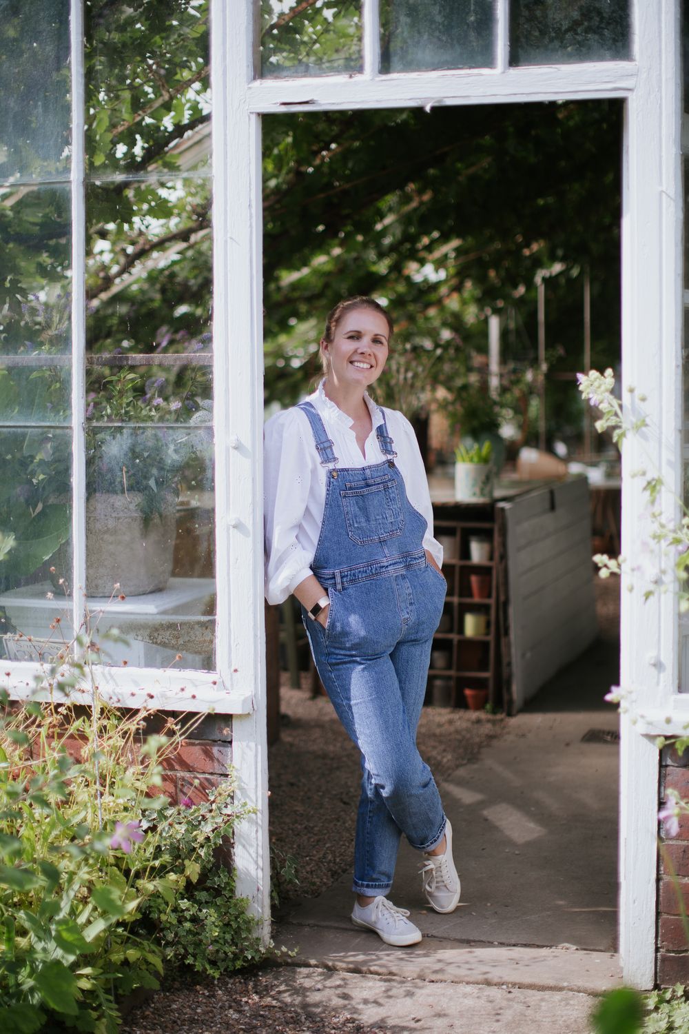 A woman leans in the doorway of a large Victorian greenhouse. She is wearing blue dungarees and white trainers