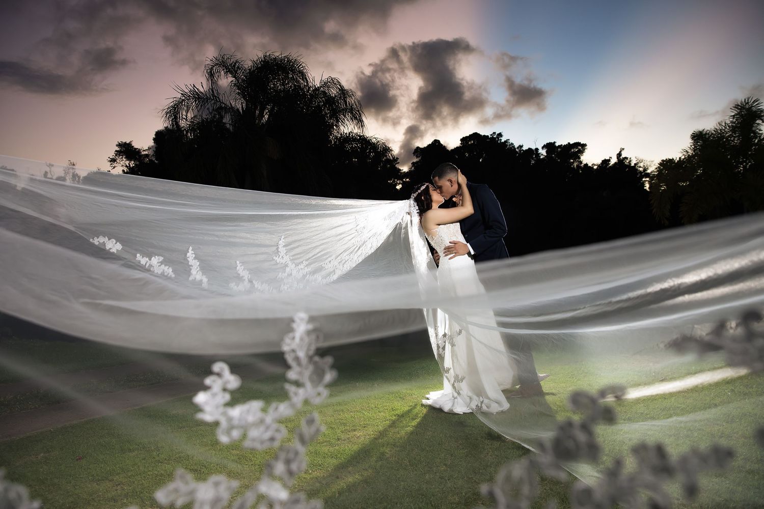 A flowing white veil catches the wind during a sunset wedding photoshoot in a garden setting.