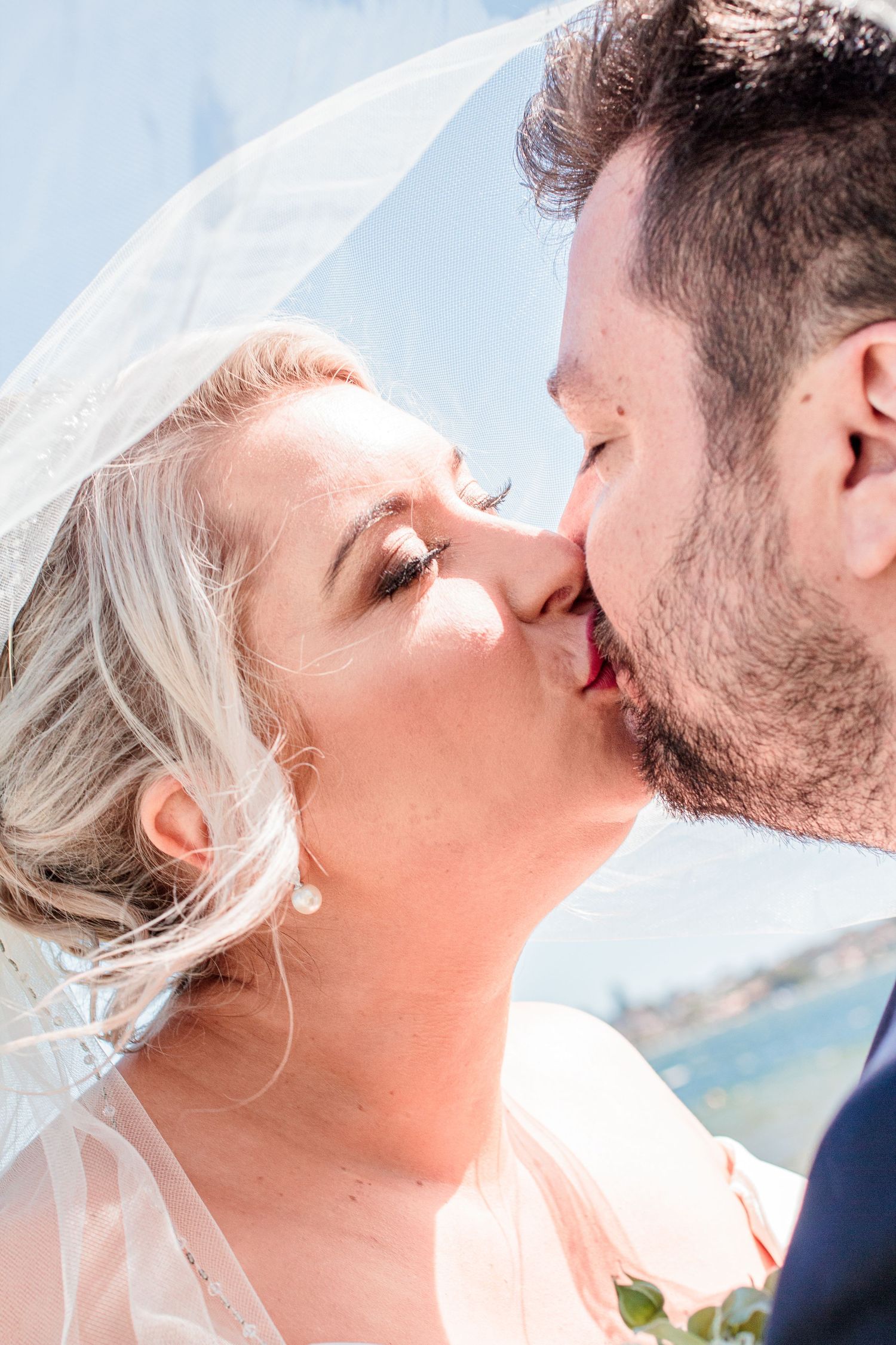 Close-up wedding portrait showing tender kiss with flowing veil against blue sky.
