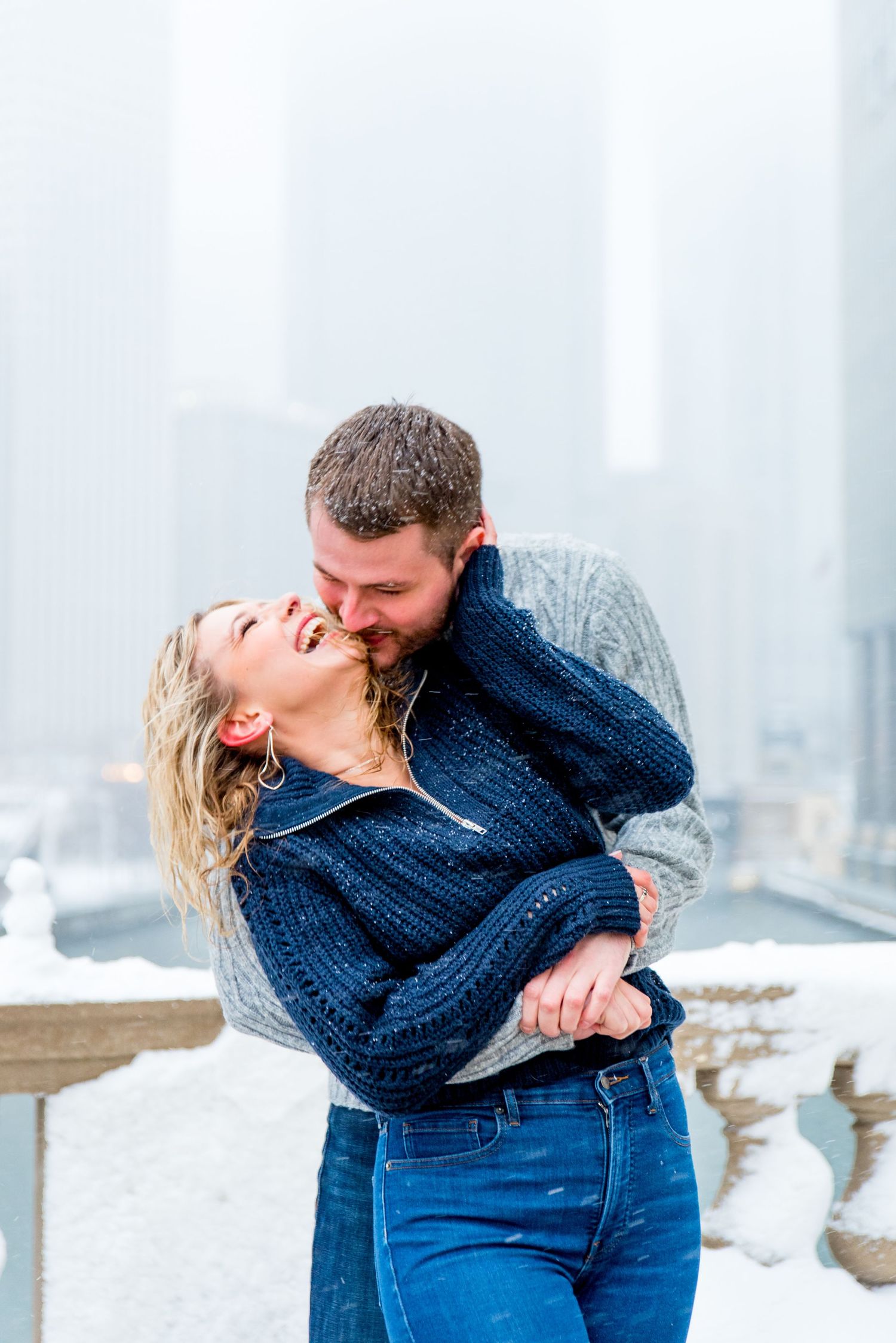 man in gray sweater hugging and kissing fiance in navy sweater at Wrigley Building for Chicago winter engagement picture