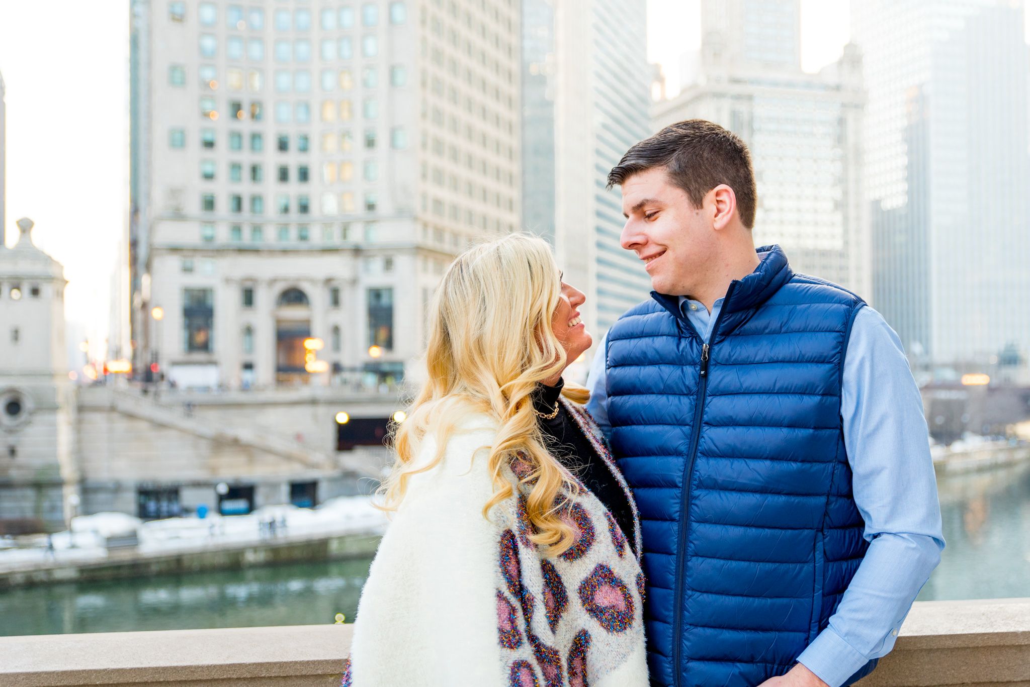blonde woman in leopard cardigan smiling at fiance in blue shirt and vest in front of Chicago River
