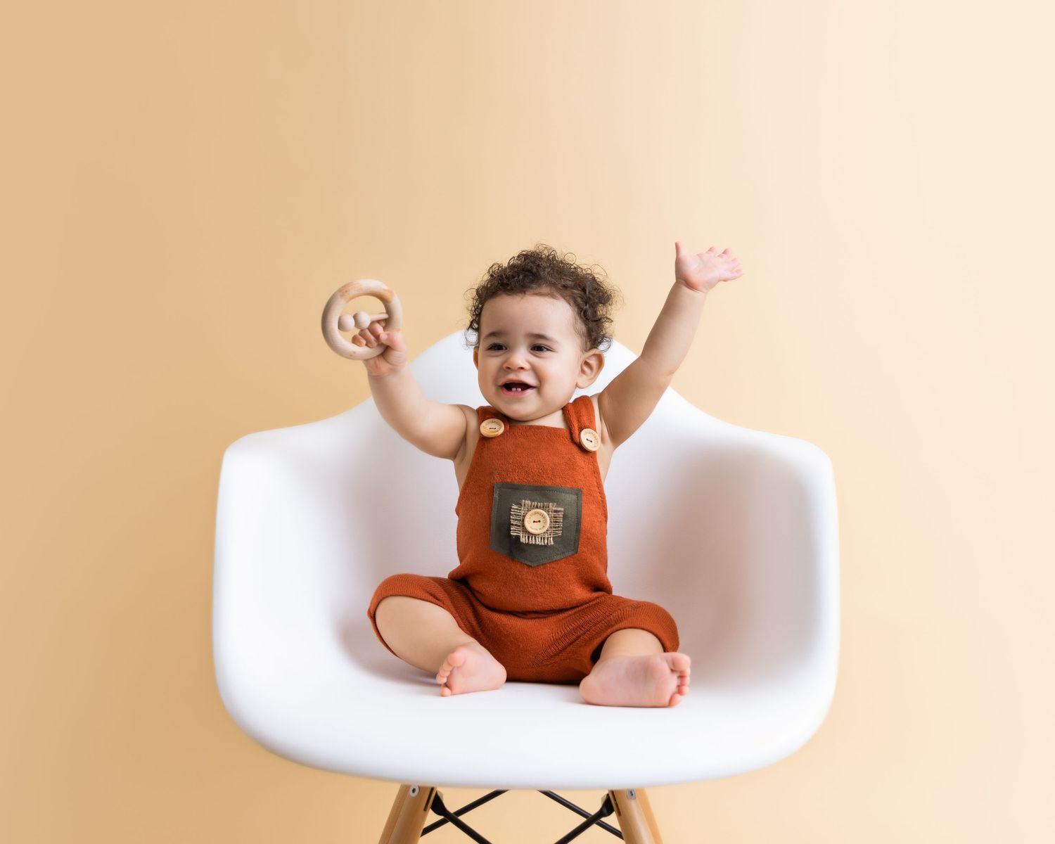 Happy baby wearing brown overalls sits in modern white Eames-style chair against beige background. Celebrating milestone