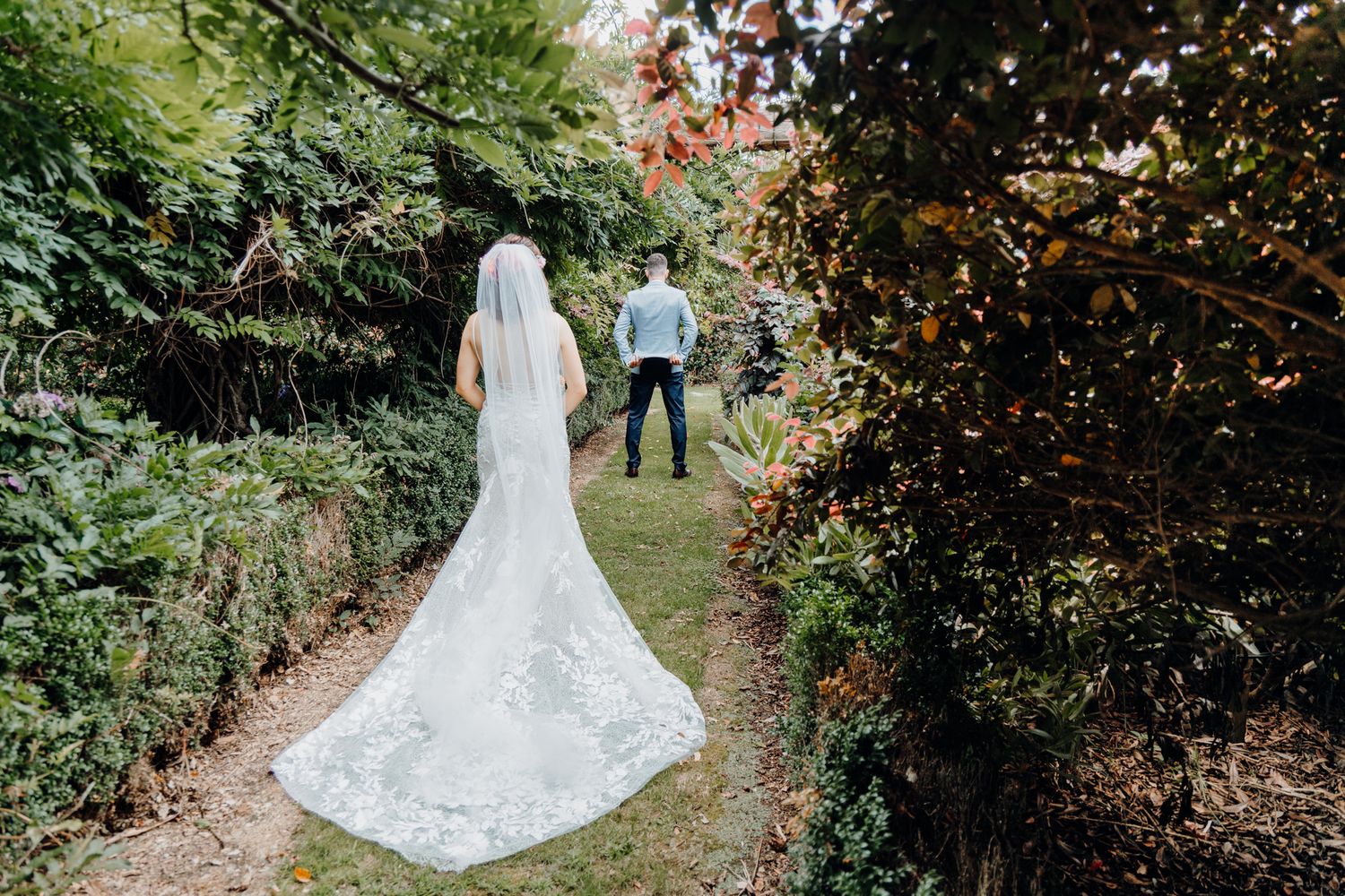 A bride in a lace wedding dress with long train poses in a garden pathway.
