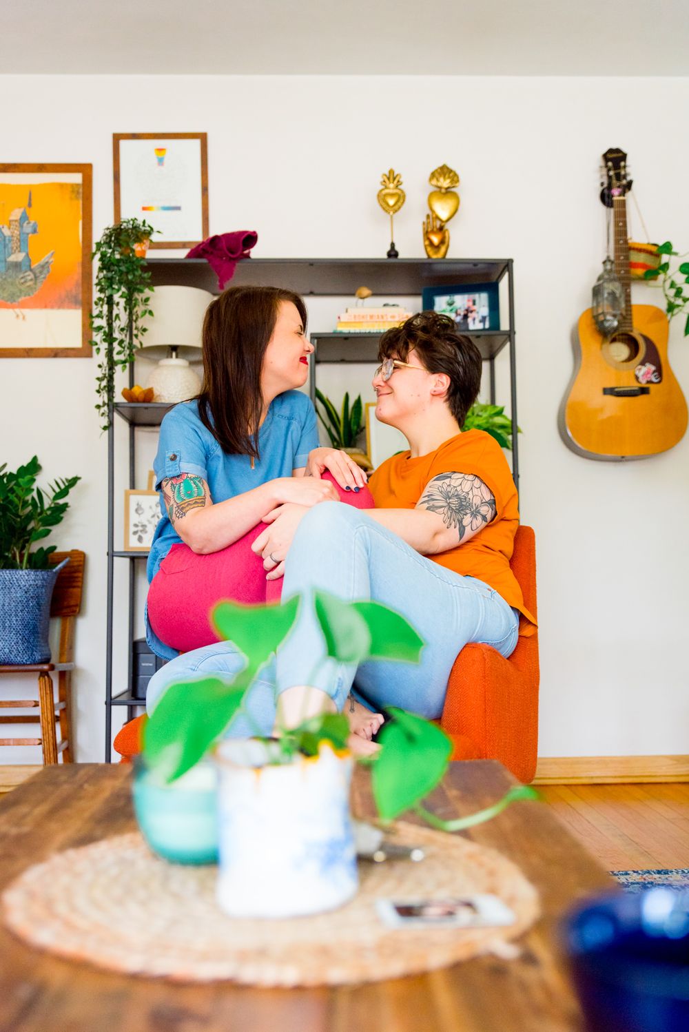 two woman in colorful clothing and colorful apartment sitting on an orange chair smiling at each other