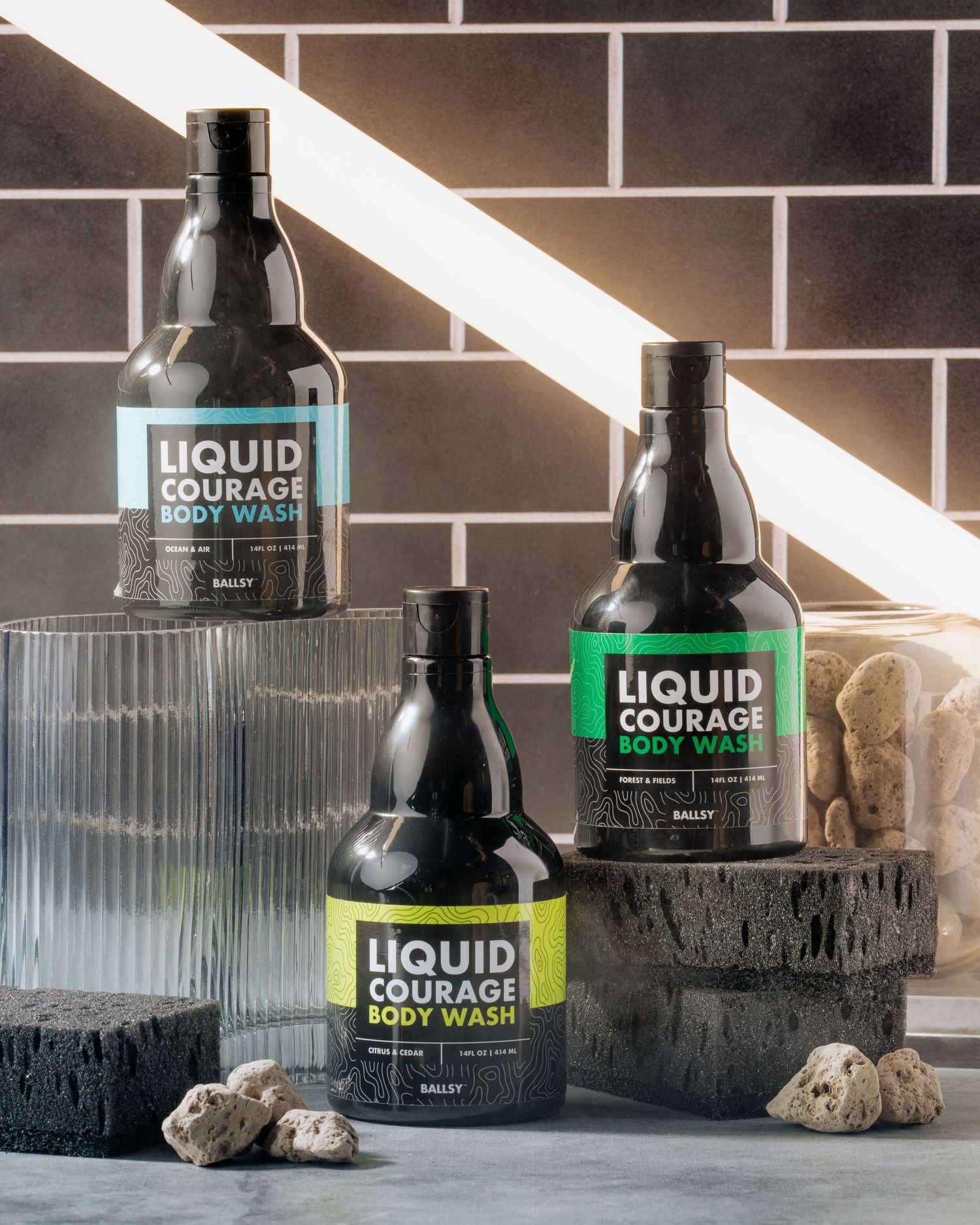 Three bottles of Liquid Courage body wash displayed with stones against brick wall.