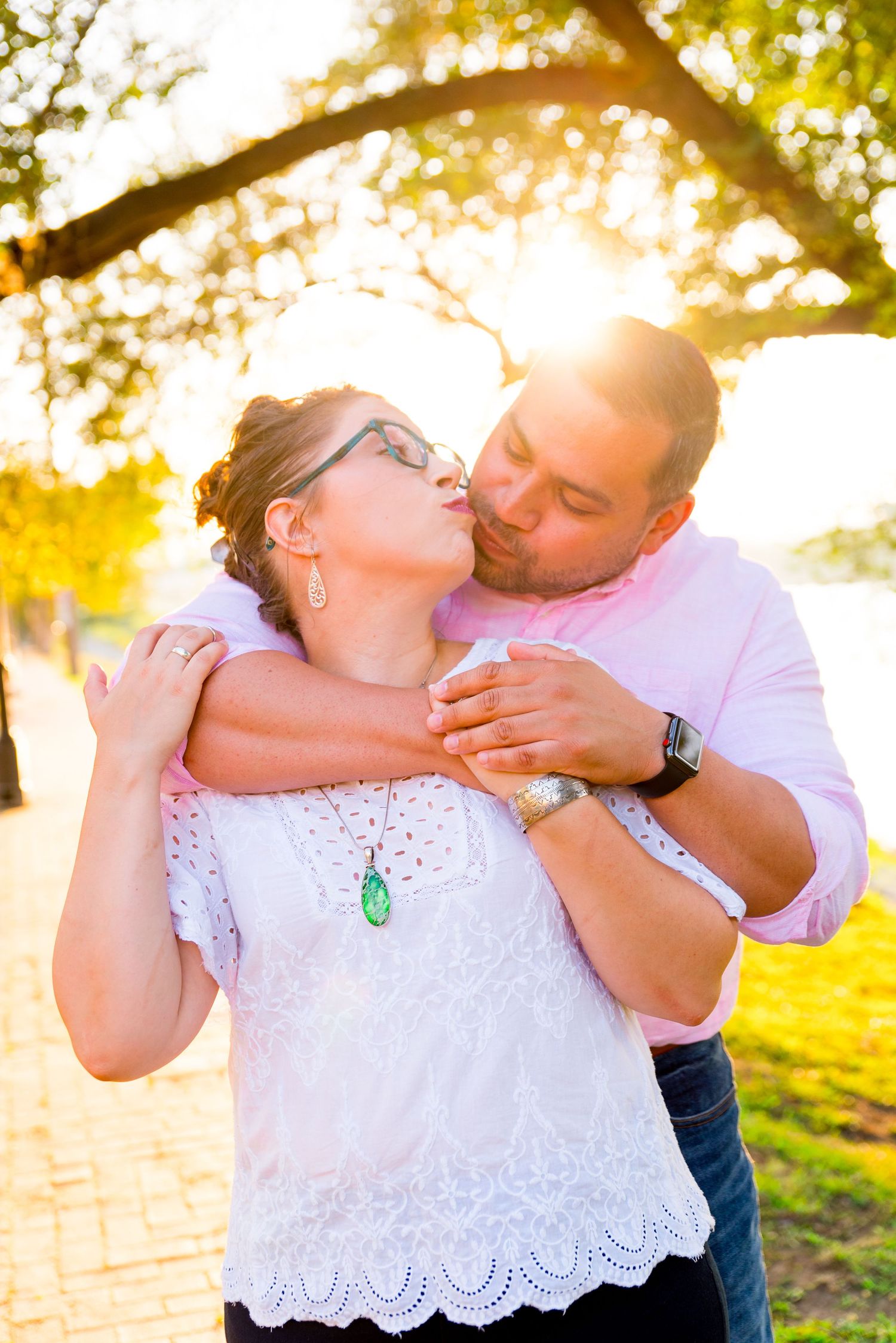 man in pink shirt hugs his fiancee in white shirt from behind and they kiss