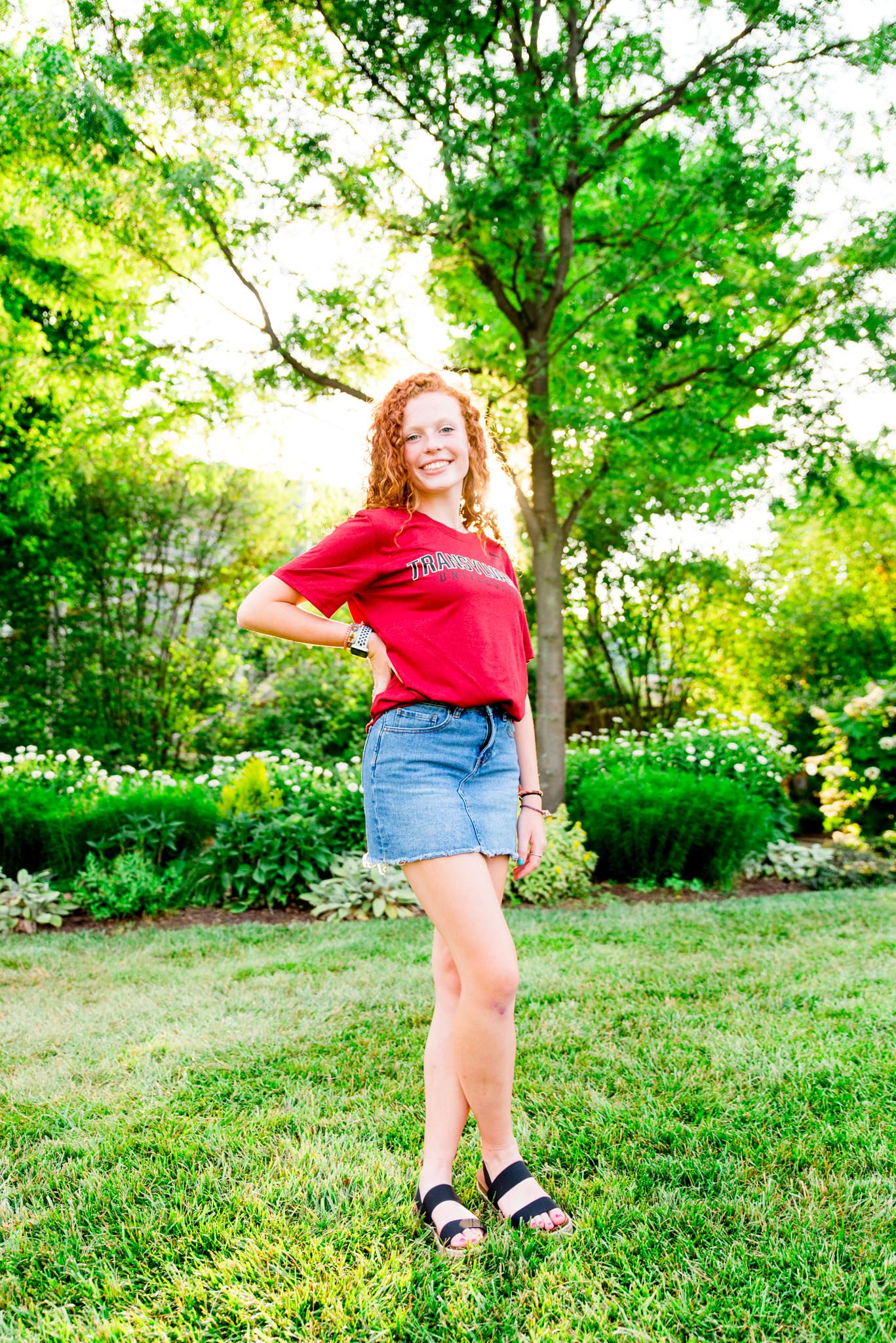 young woman in red tshirt in front of trees at Smale Park at sunset with one hand on hips