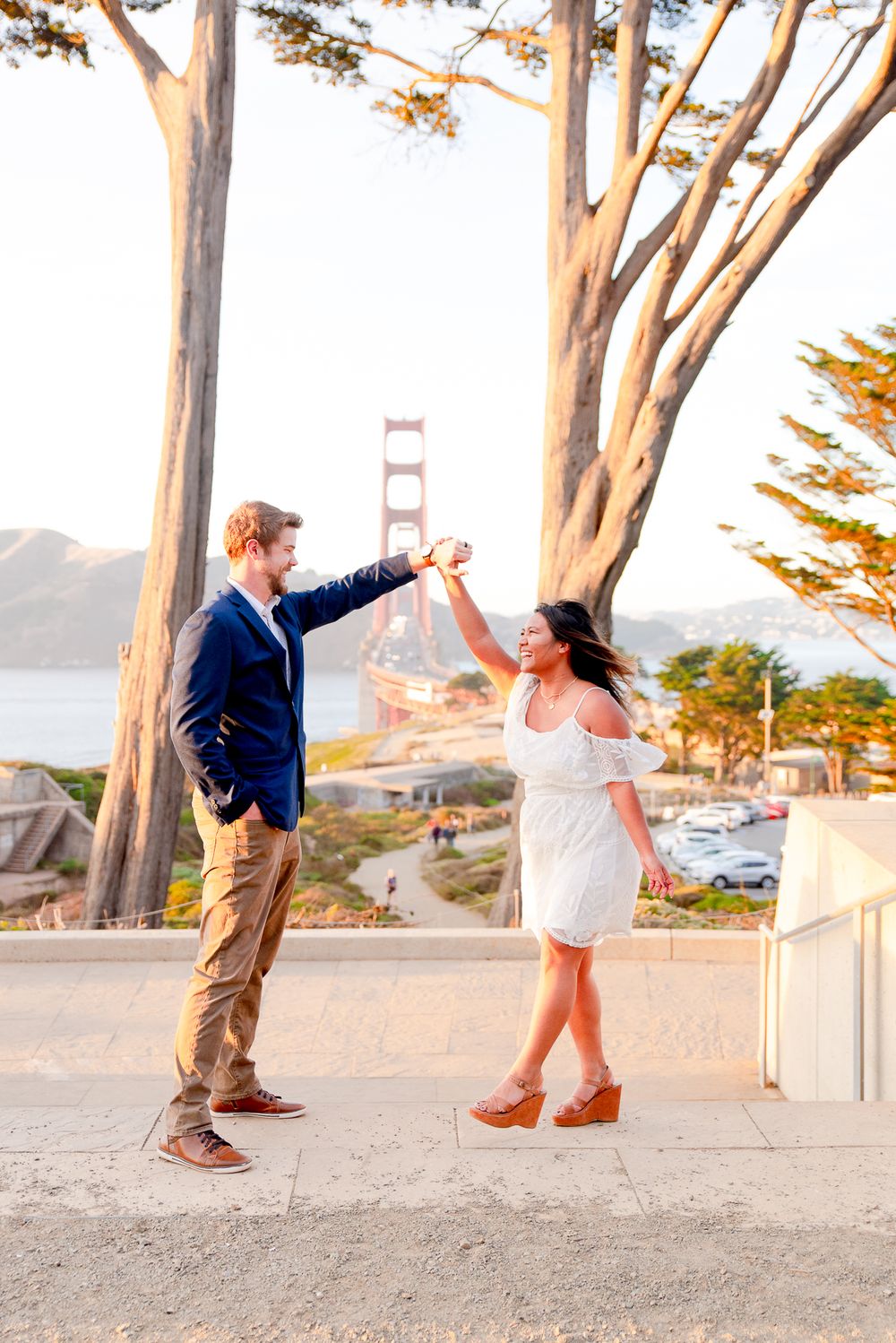 man in navy suit twirling woman in white dress at sunset in hills in front of Golden Gate Bridge in background
