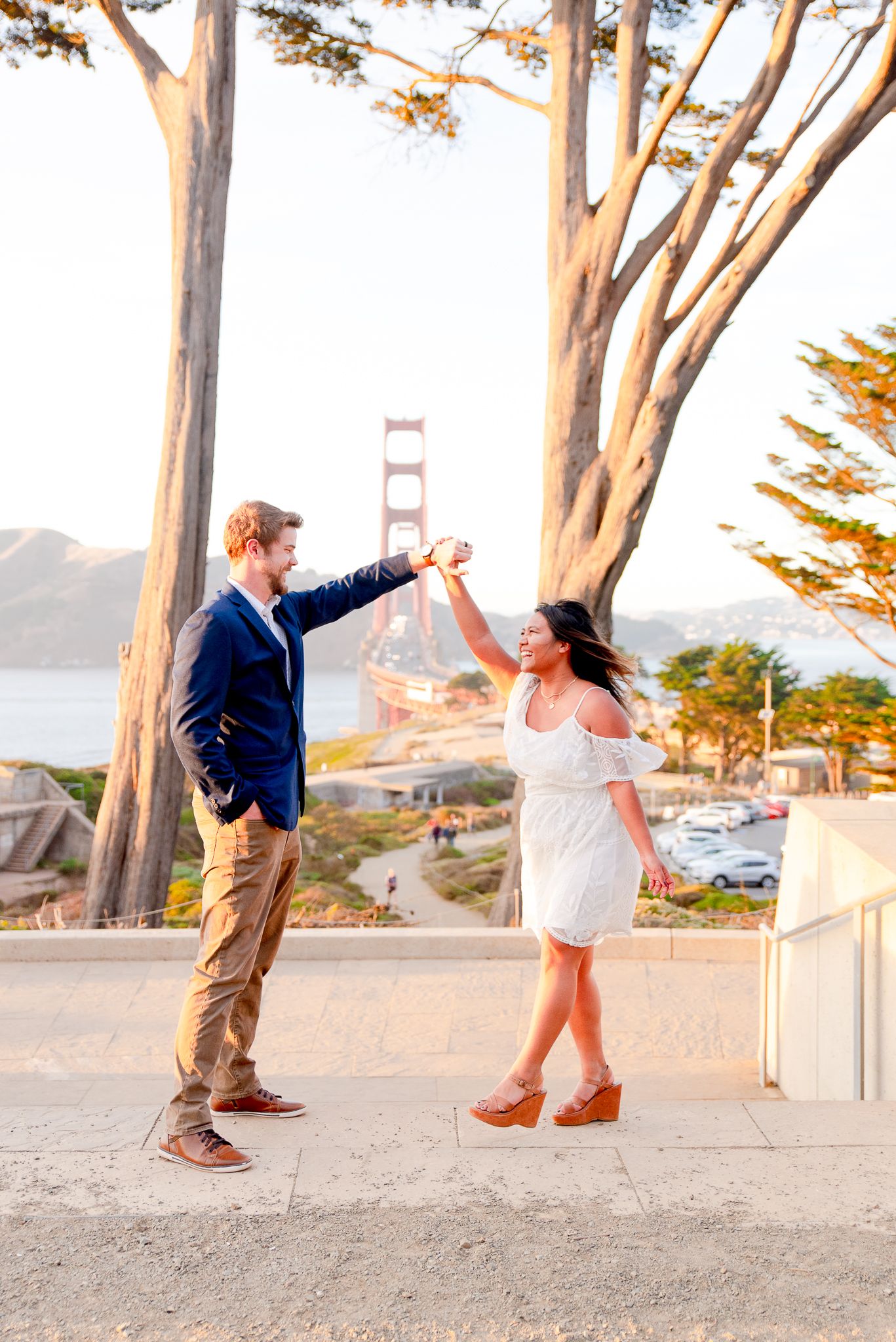 man in navy suit twirling woman in white dress at sunset in hills in front of Golden Gate Bridge in background