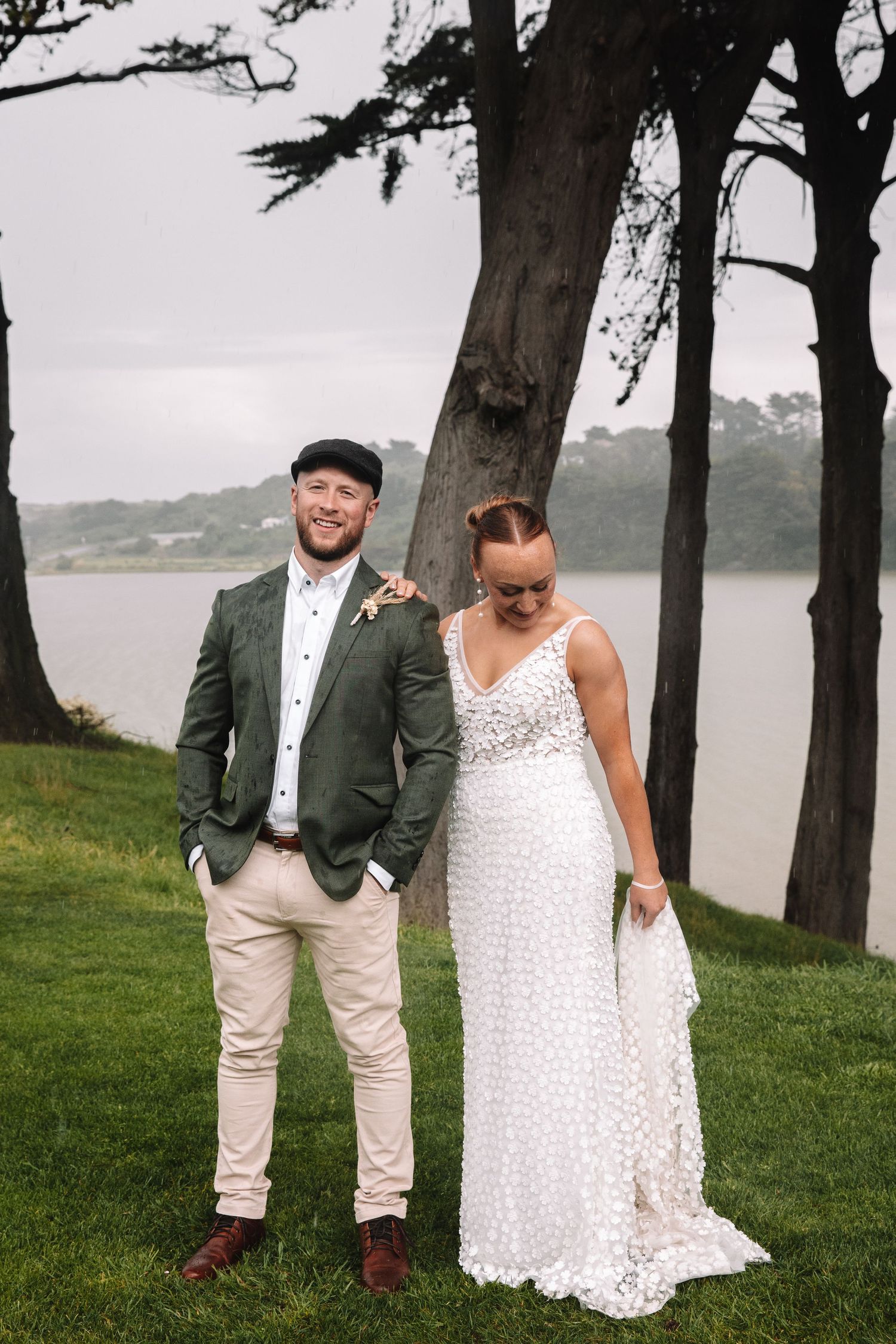Newlyweds posing by a lake with trees, bride in lace dress and groom in casual suit.