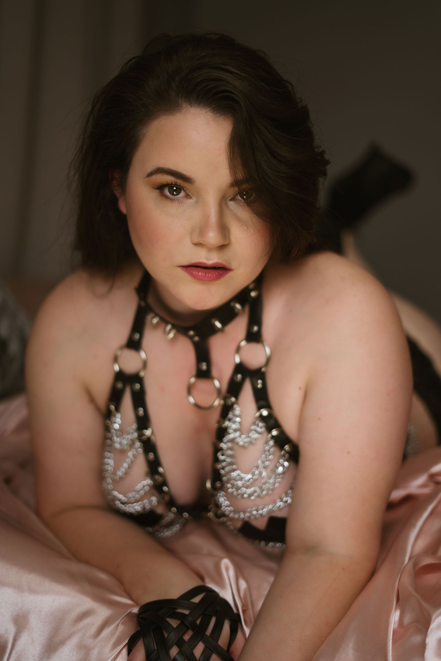 A woman in a leather and chain harness lies on her front, facing the camera head on