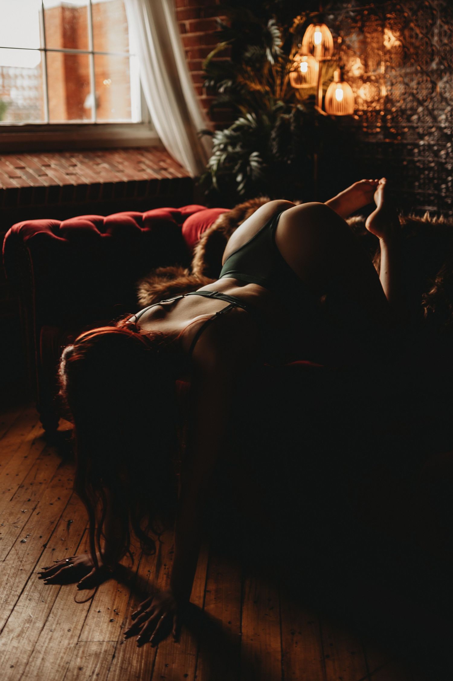 Figure lounging on red velvet sofa against brick wall with hanging pendant lights.