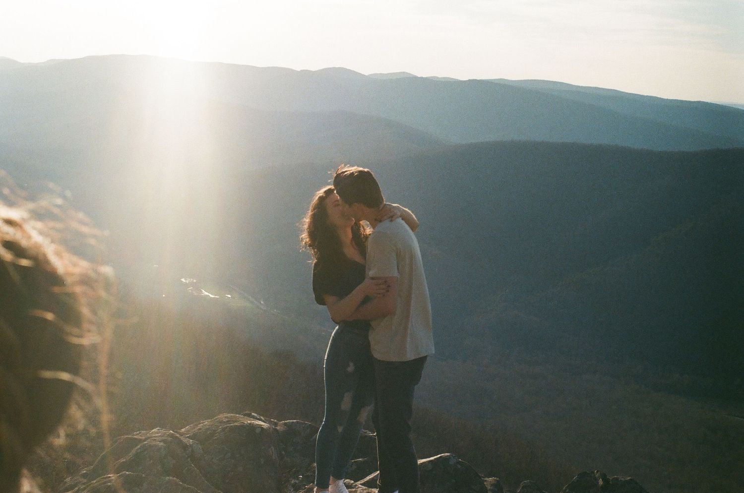 A couple embraces on a mountaintop overlook with sunlight streaming through the scenic landscape.