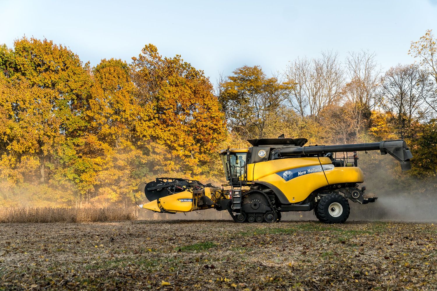 Yellow combine harvester working in autumn field with golden foliage in background.
