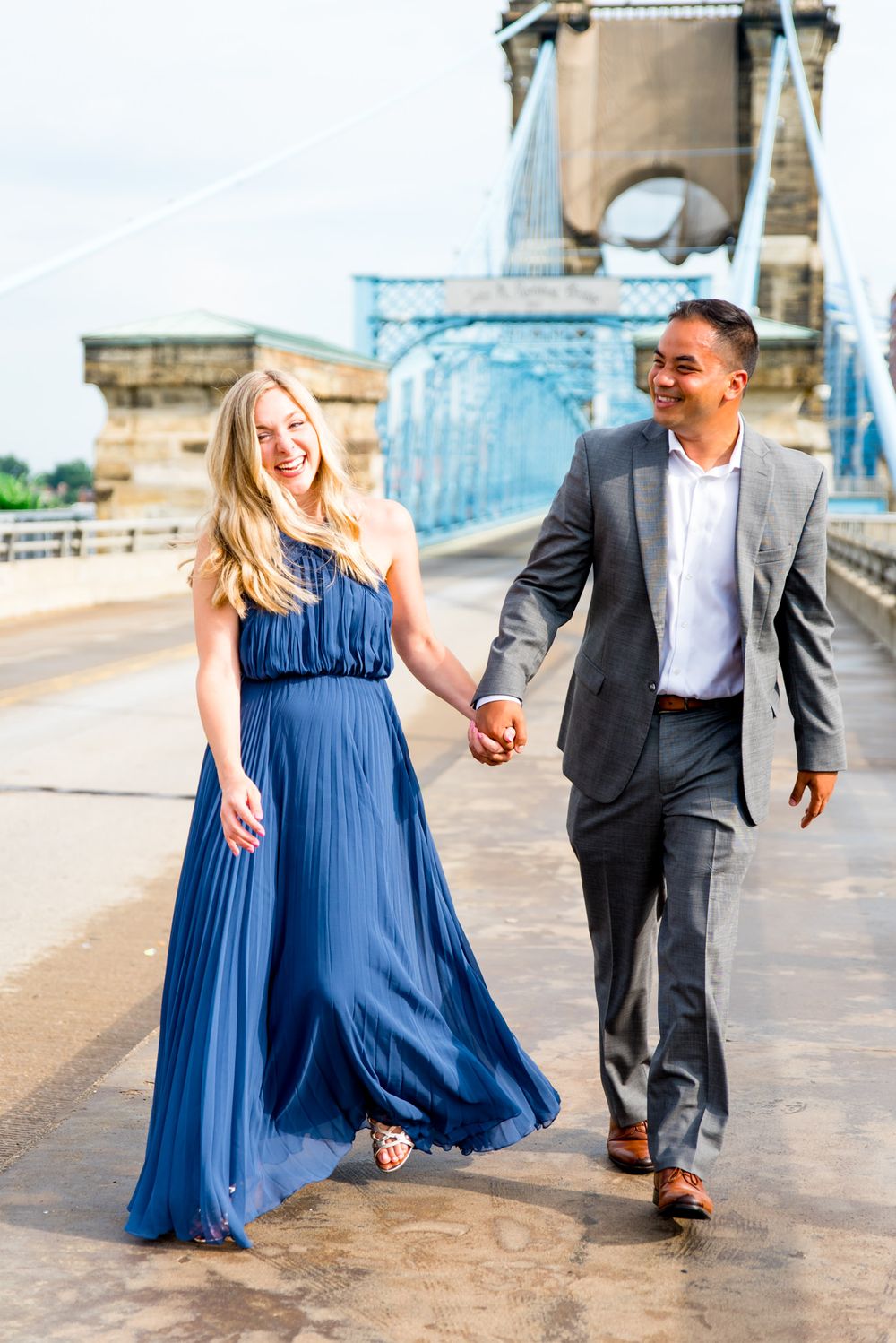 man in grey suit holds hand of blonde woman in blue dress as they walk near the Roebling Bridge for engagement pictures