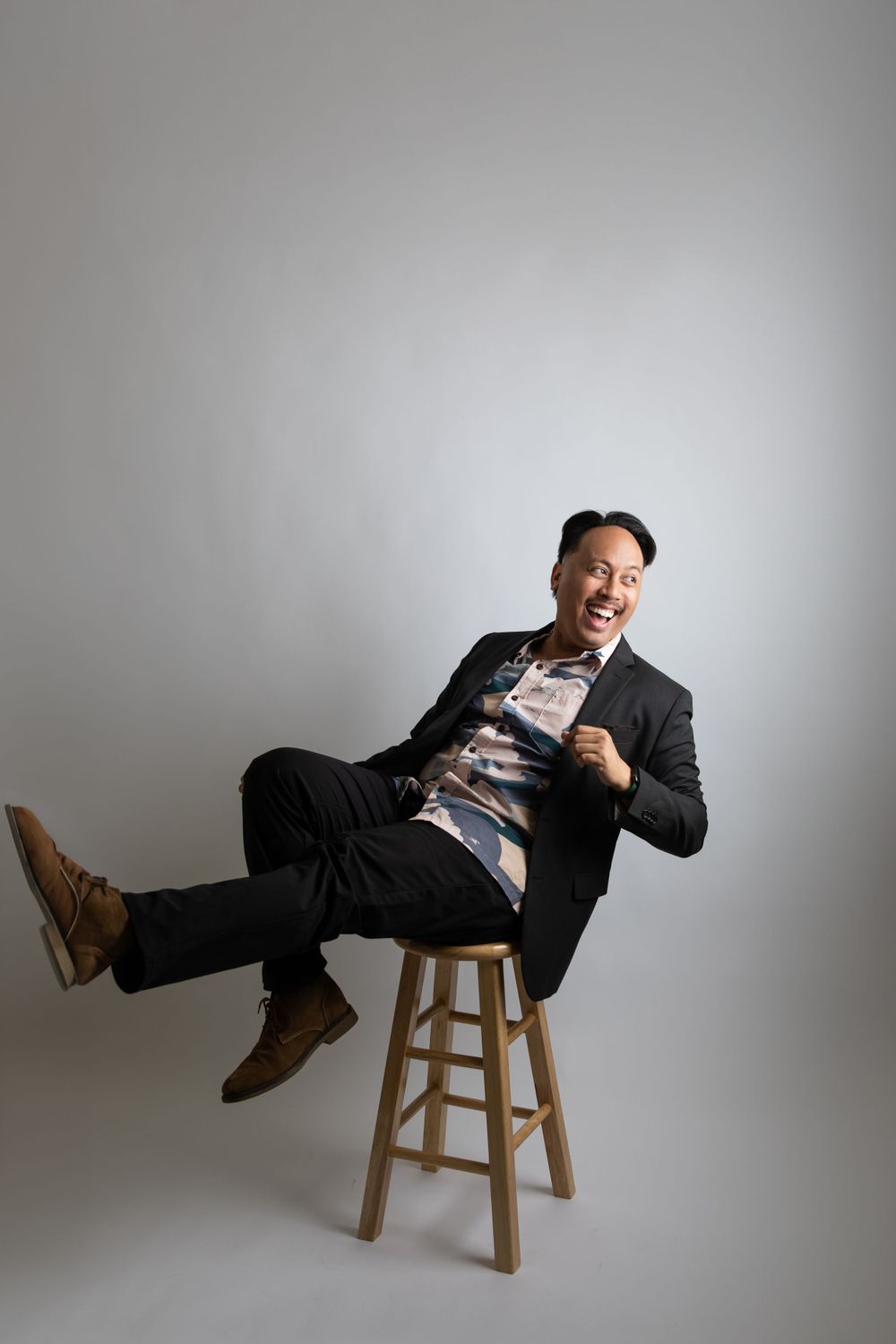 Professional portrait of someone playfully leaning back on a wooden stool against a gray background.