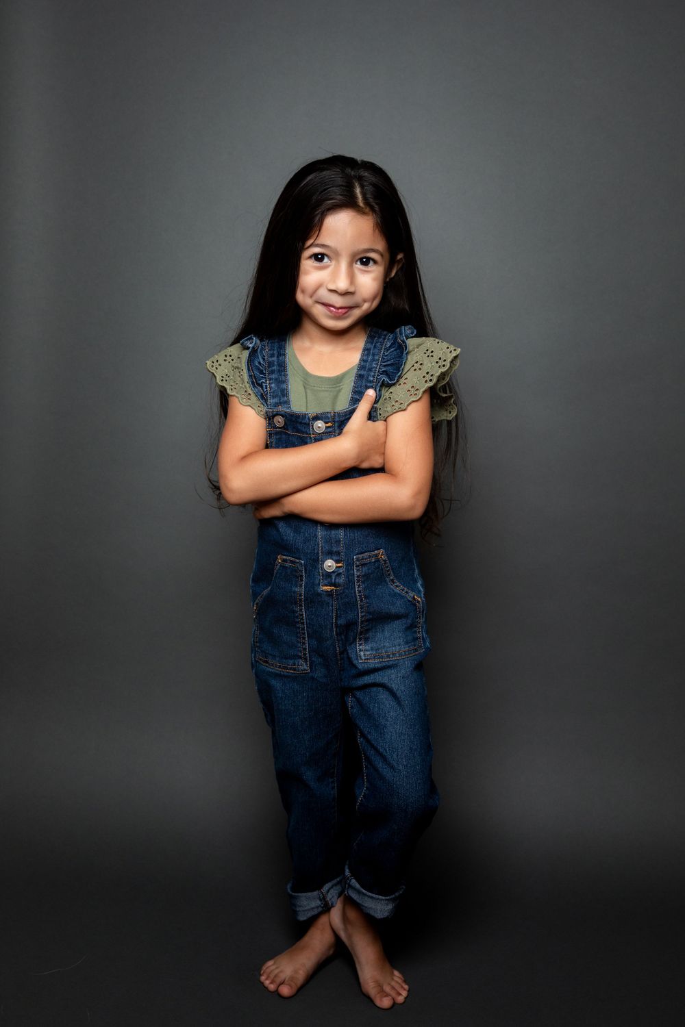 Young child wearing denim overalls poses confidently against dark background.