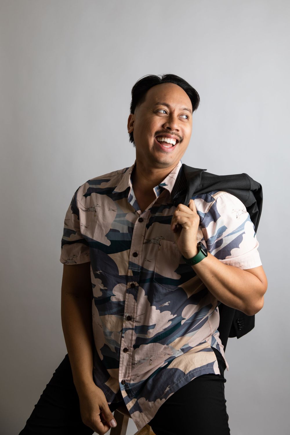 Casual studio portrait of someone in a camouflage shirt with a black jacket over shoulder.