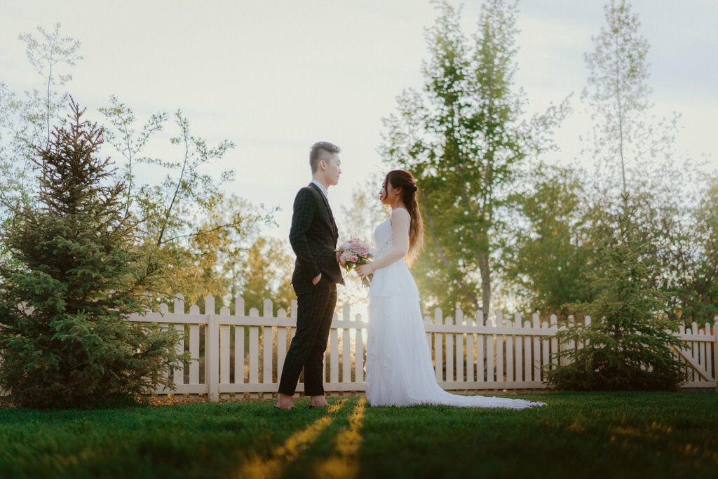 A romantic sunset wedding portrait with a white fence and trees creating a dreamy backdrop.