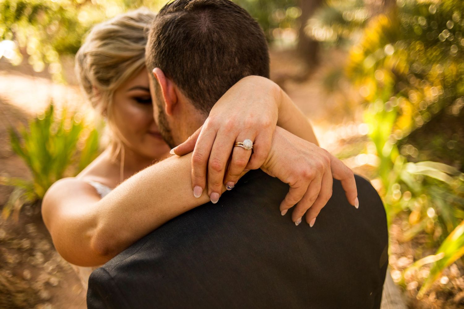 Close-up of bride's hands with wedding ring embracing groom in golden sunlight.