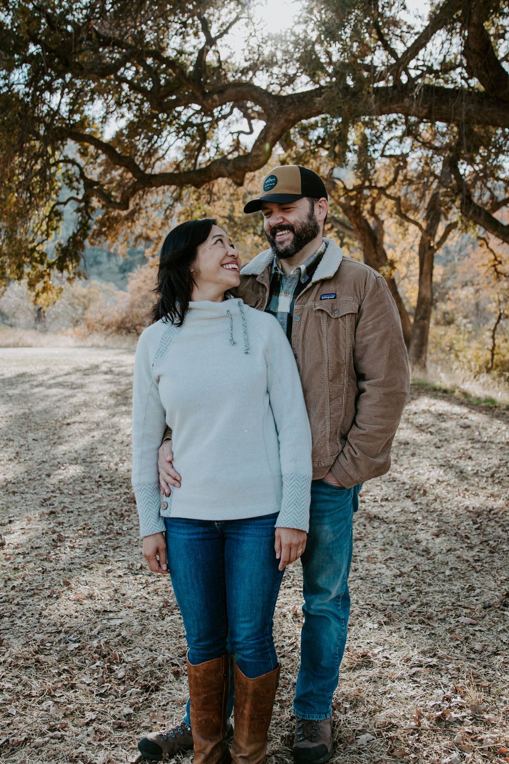 Couple in casual winter wear standing together on a dirt path under oak trees.