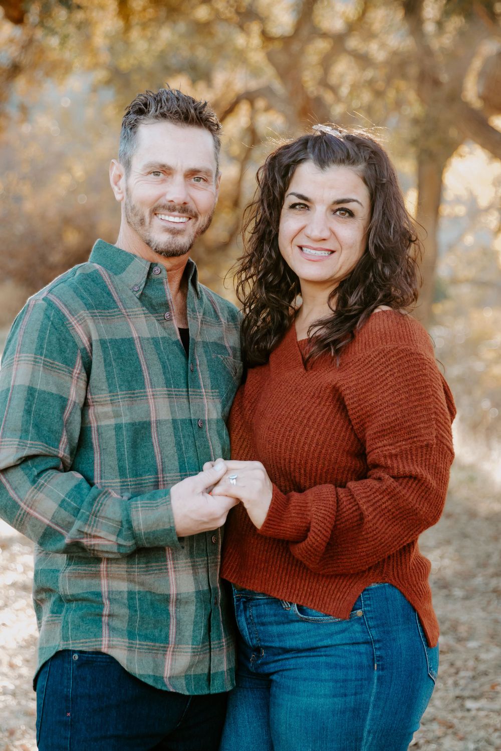 A couple in casual fall attire smiling together outdoors with a plaid shirt and rust sweater in autumn lighting.