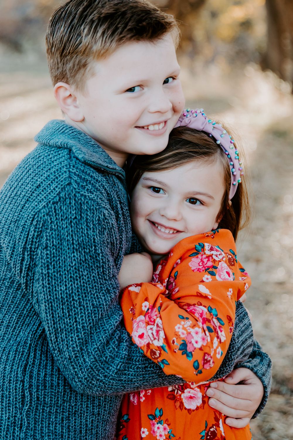 Two children embracing while wearing knit sweaters and floral dress against a blurred natural background.