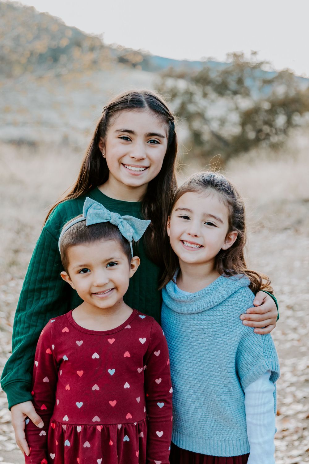 Three children in sweaters posing together for a portrait with mountains in background.