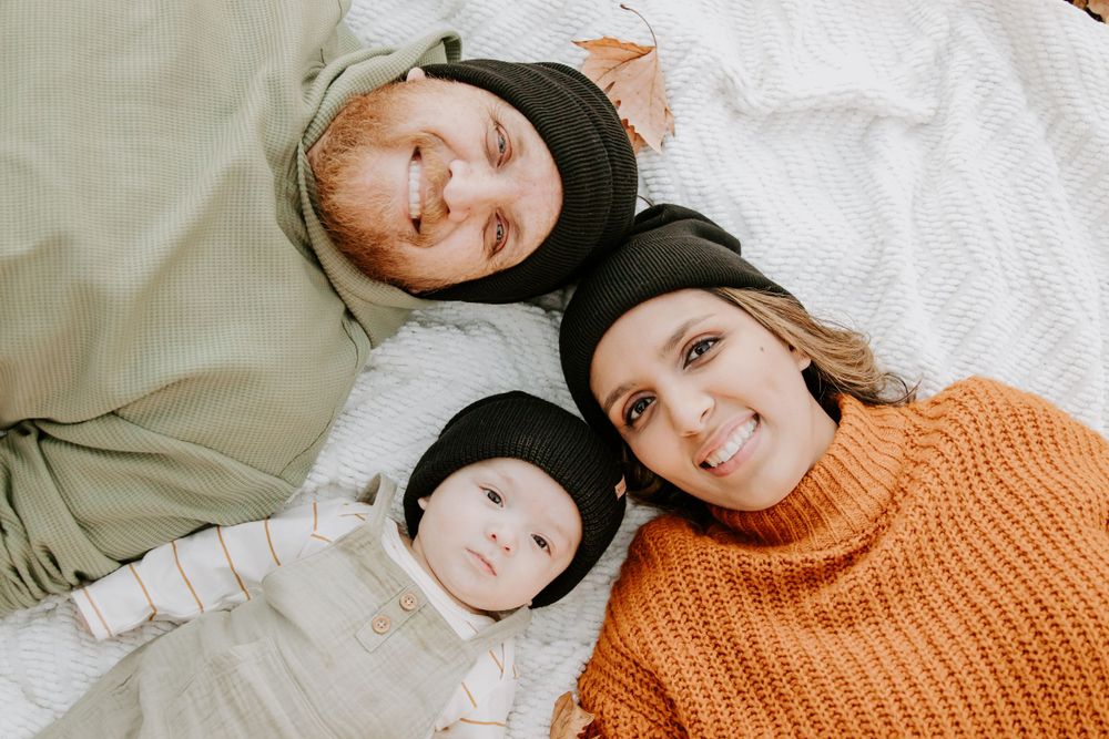 A warm cozy family portrait of three people in winter clothing laying together on a white blanket.
