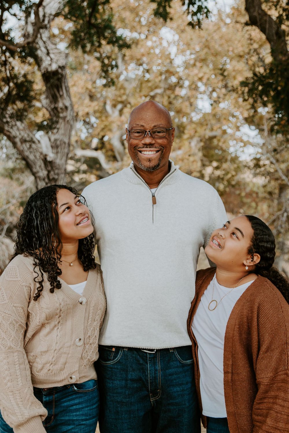 Three people in casual sweaters pose together outdoors with autumn foliage in background.