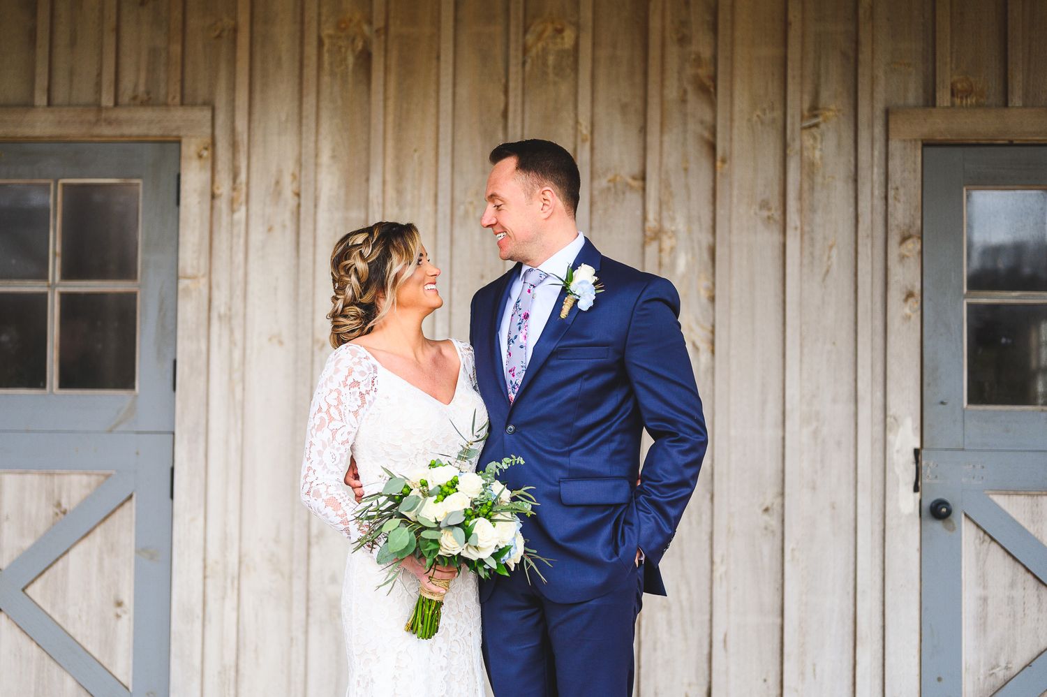bride and groom smiling at each other in front of rustic barn