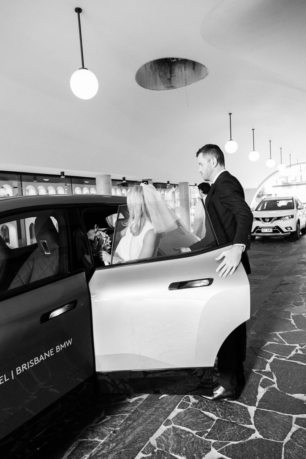 Groom helps bride into car in black and white wedding photo.