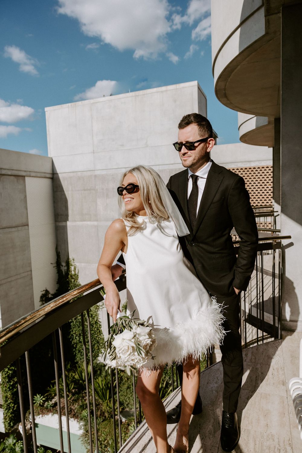 Modern wedding couple in sunglasses pose against curved architectural backdrop.