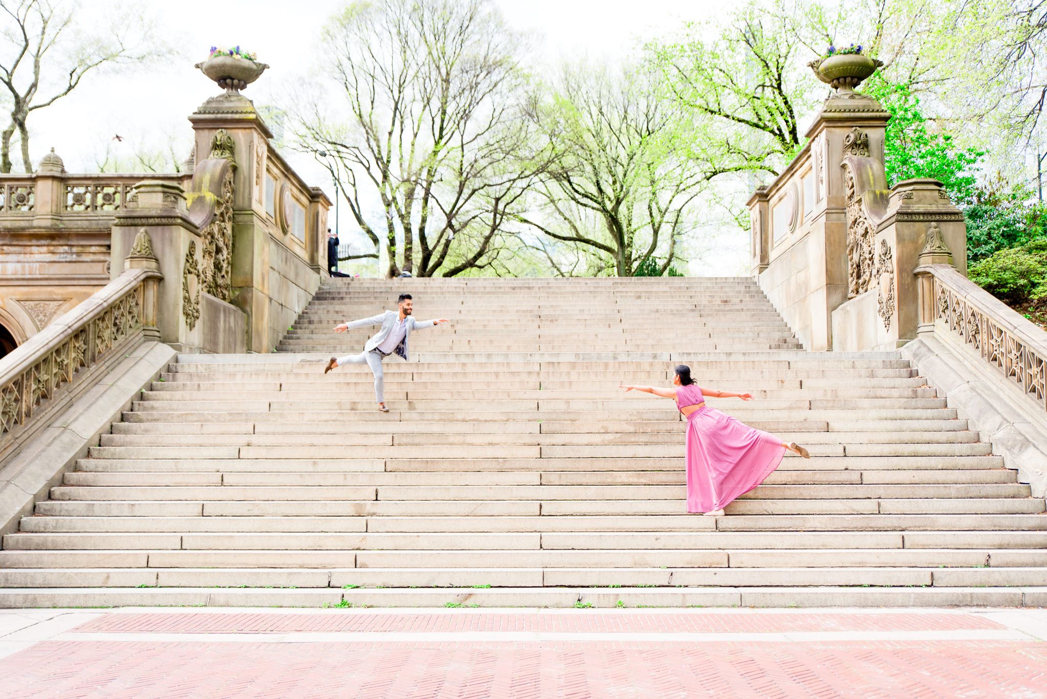 Woman in pink dress and man in gray suit dancing on steps to Bethesda Terrace Central Park for spring engagement photos