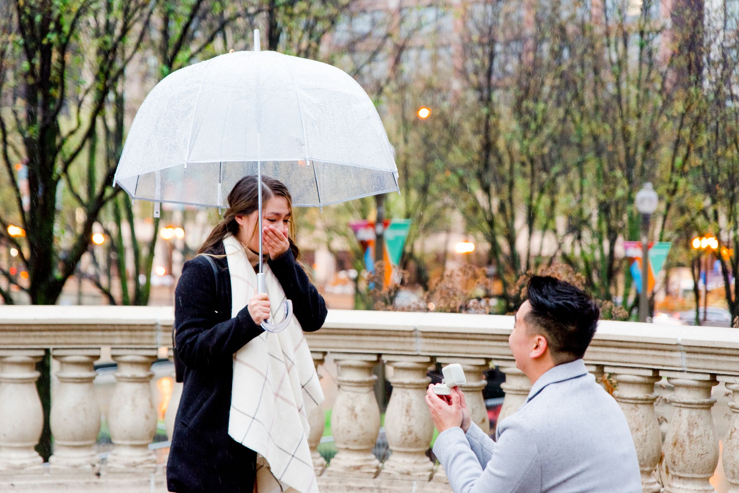 man in grey coat kneeling proposing to woman in black coat and clear umbrella in front of Millennium Park Christmas Tree