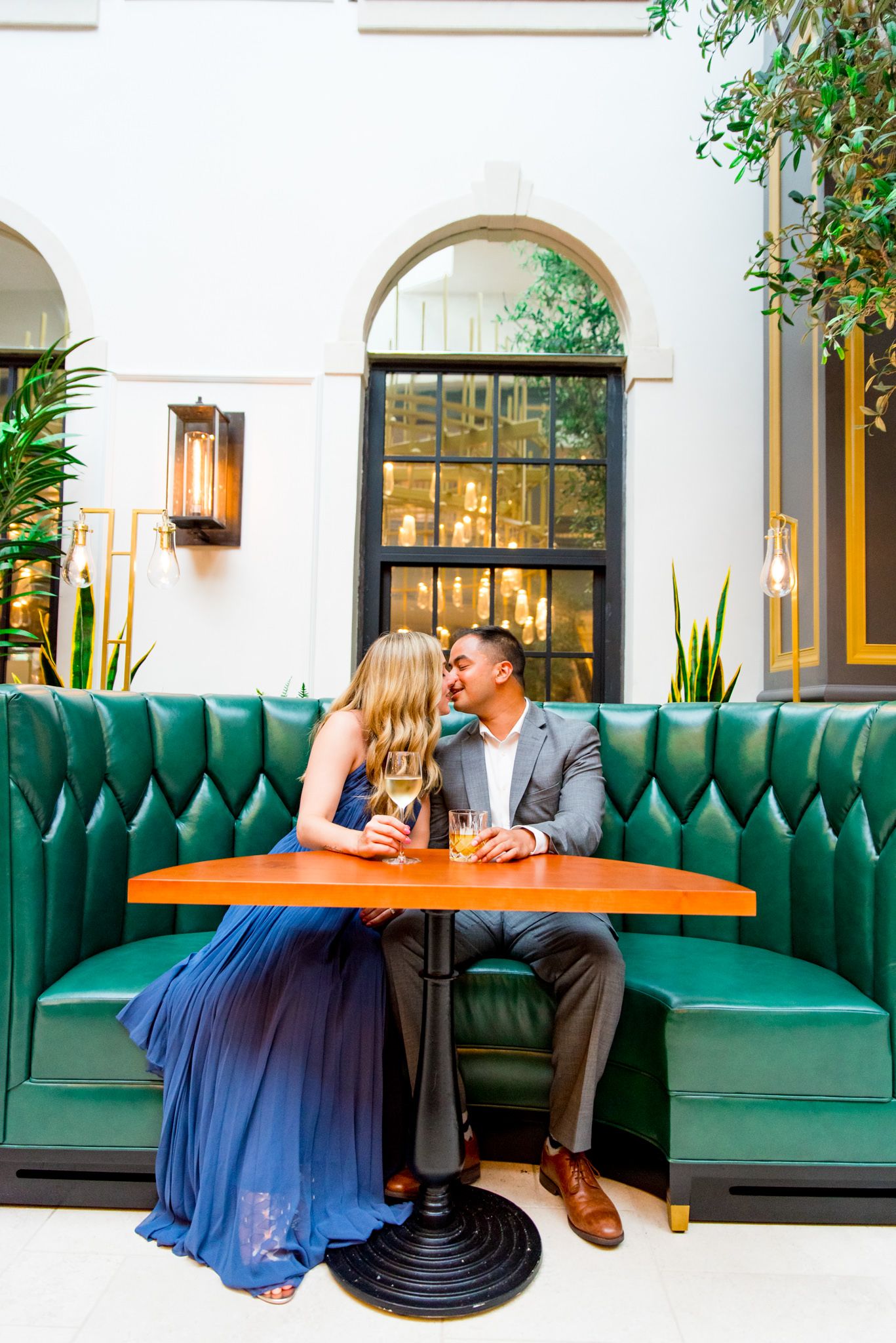 man in grey suit and woman in blue dress holding a glass of white wine kiss in green booth at Lytle Park Hotel