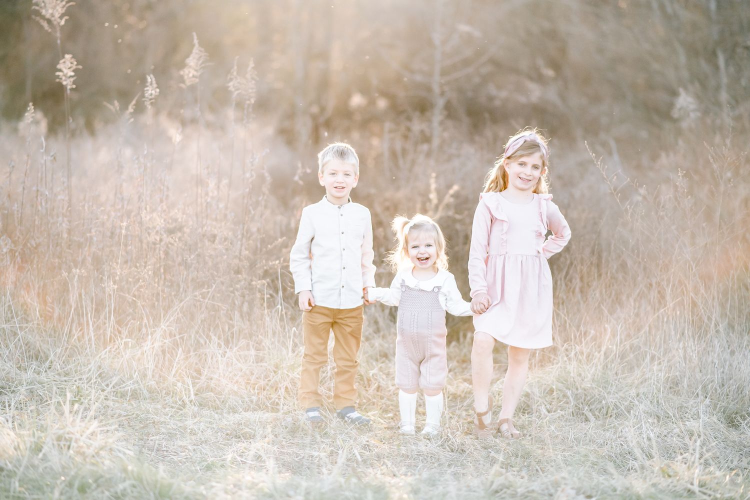 Three siblings hold hands in a field with sun flare around them